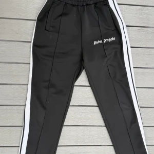 Palm angels byxor - Svarta byxor från Palm Angels med vita sidoränder och logga på vänster ben. Byxorna har elastisk midja och raka ben, tillverkade i polyester för en sportig look.PRISET är ej hugget i sten