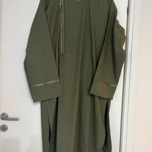 Olivgrön afghansk traditionell kläder för män.  - Säljer en olivgrön afghanskt attire med raka långa ärmar och dekorativa detaljer längs halsringningen och ärmsluten. Den har en enkel och stilren design, perfekt för dig som gillar minimalistiskt mode. Kommer med matchande byxor. Kommer direkt från Afghanistan. Köpt för 1400 kr denna året.
