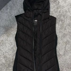 Svart dunväst med huva från Zara - Säljer en svart dunväst från Zara med huva och dragsko. Västen har ett stilrent, vadderat mönster och är perfekt att bära över en hoodie eller tröja. Passar dig som gillar en sportig och trendig look.