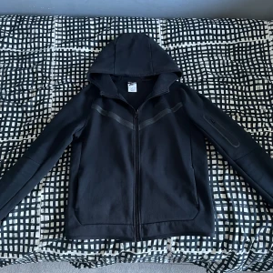 Nike tech - Säljer en svart kofta från Nike med dragkedja framtill och huva. Koftan har en diskret ficka med dragkedja på ärmen och stilrena detaljer. Koftan har inga fel. Vid snabb affär kan priset diskuteras. Vid funderingar är det bara att skriva.