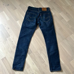 Mörkblåa Levis jeans - Mörkblå jeans från Levi's modell 512 med klassiska fem fickor och raka ben. Jeansen har kontrastsömmar, läderpatch bak i midjan och Levi's röda etikett på bakfickan. Perfekta för en stilren vardagslook.