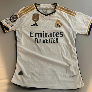 Real Madrid hemmatröja 2023/24 Camavinga #12 - Säljer en vit Real Madrid hemmatröja från säsongen 2023/24 med Camavinga och nummer 12 på ryggen. Tröjan har guld- och svarta detaljer, broderat klubbmärke, FIFA World Champions-märke och Champions League-patch. Tillverkad i lätt funktionsmaterial. Spelarversion, äkta, nyskick.