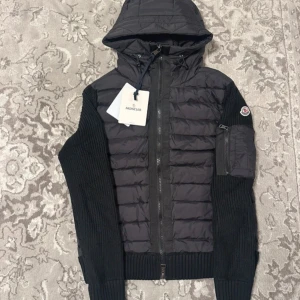 Svart hybridjacka med huva från Moncler - Säljer en svart Moncler-jacka med quiltad front och huva. Ärmarna är stickade och har en ficka med Moncler-logga på vänster arm. Jackan har dragkedja framtill och ribbade muddar.