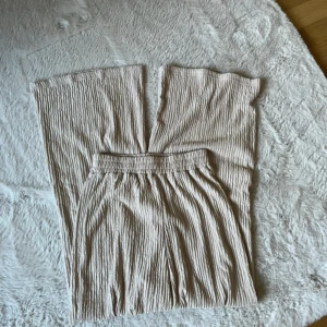 Beige plisserade vida byxor med matchande linnetopp - Säljer ett par beige plisserade byxor med resår i midjan och vida ben. Byxorna är i ett mjukt, luftigt material och har en avslappnad passform. Matchande ärmlös linnetopp ingår, perfekt för en chill och stilren look. Aldrig använda.
