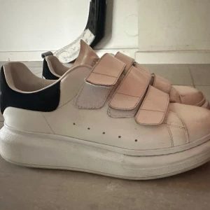 Vita sneakers från Alexander McQueen med kardborreband - Säljer ett par vita sneakers från Alexander McQueen med tre breda kardborreband och svart detalj på hälen. Superfina nu till sommaren! Lite smutsiga men annars som nya!