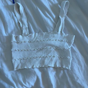 Vit ribbad croppad linnetopp - Säljer en vit ribbad croppad linnetopp med breda axelband och vågiga volangdetaljer. Toppen har en stretchig passform och är perfekt sommartopp