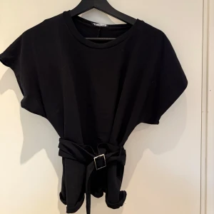 Svart topp med bälte från Zara - Snygg svart topp från Zara med rund halsringning och korta ärmar. Toppen har ett brett tygbälte i midjan med silverfärgat spänne som ger en markerad siluett. Perfekt att styla till jeans eller kjol.