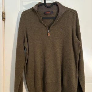 Olivgrön stickad tröja med dragkedja från Morris - Säljer en olivgrön stickad tröja från Morris med halv dragkedja och hög krage. Tröjan har långa ärmar och diskret broderad logga på bröstet. Perfekt för lager-på-lager och snygg till jeans eller chinos. Liten i storlek passar S-L