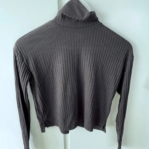 Svart ribbad polotröja från Divided - Säljer en svart ribbad polotröja från Divided. Tröjan är croppad och har långa ärmar samt en hög polokrage. Perfekt för lager på lager och enkel att matcha med olika outfits.