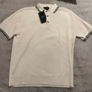 Vit piké från Massimo Dutti - Säljer en stilren vit pikétröja från Massimo Dutti med klassisk krage och svarta ränder på krage och ärmslut. Tröjan har knappar framtill och är tillverkad i bomull. Perfekt för en clean och fräsch look.