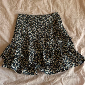 Blommig volangkjol - Supersöt kort kjol med blå botten och små blommor. Kjolen har volangdetaljer och passar perfekt till varmare dagar nu i sommar. Bekväm och lätt att matcha med olika toppar.