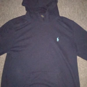 Mörkblå hoodie från Polo Ralph Lauren - Säljer en mörkblå hoodie från Polo Ralph Lauren med klassisk broderad logga på bröstet i ljusblått. Tröjan har huva och magficka, perfekt för en avslappnad stil. Custom fit-modell. Kontakta gärna mig ifall du skulle ha några funderingar, pris kan diskuteras.