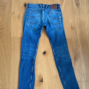 Replay Anbass slim fit jeans - Säljer ett par blå Replay Anbass jeans med slim fit. Klassisk femficksmodell med snygga slitningar och Replay-logga på bakfickan. Jeansen har dragkedjegylf och detaljer i rött vid fickan framtill.