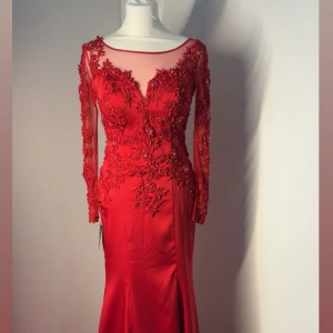 Röd långklänning med spets och öppen rygg - Elegant röd långklänning med figurnära passform. Klänningen har långärmade ärmar och en vacker öppen rygg med knappstängning. Perfekt för bal eller festliga tillfällen. Orginal priset är 2500kr