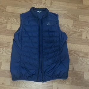 Mörkblå vadderad väst från 01TP - Säljer en mörkblå vadderad väst från 01TP i storlek L (44/46). Västen har dragkedja framtill, quiltad framsida och en praktisk ficka med dragkedja på ryggen. Perfekt för lager-på-lager under kyliga dagar.