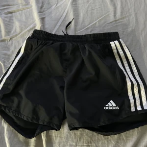Svarta träningsshorts från Adidas - Svarta träningsshorts från Adidas med klassiska vita ränder längs sidorna och Adidas-logga framtill. Shortsen har elastisk midja och är tillverkade i ett lätt syntetmaterial, perfekta för sport och träning.