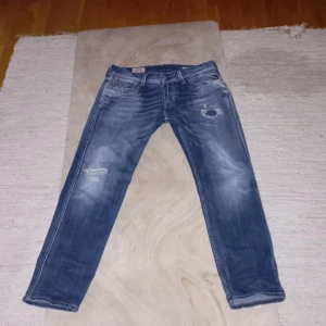 Replay Anbass jeans med slitningar och - Säljer ett par blå Replay Anbass jeans med snygga slitningar och hål på framsidan. Byxorna har en unik dragkedjedetalj på bakfickan och klassisk femficksdesign. Perfekta för dig som gillar en avslappnad stil