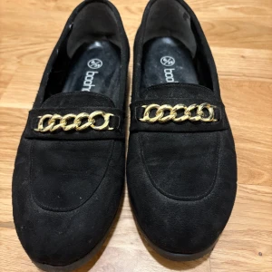 Svarta loafers med guldkedja - Snygga svarta loafers från Boohoo med en elegant guldkedja på ovansidan. Skorna är tillverkade i mocka och har en klassisk design som passar perfekt för en stilren look.