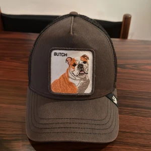 Grå/svart keps med broderad bulldog - Snygg grå keps från Goorin Bros med ett broderat märke av en bulldog på framsidan. Kepsen har ett nätmaterial baktill för extra ventilation och en justerbar rem för perfekt passform.