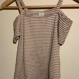 Randig topp från Lindex - Säljer en randig topp från Lindex i beige och svart. Toppen har en offshoulder-design med smala axelband och korta ärmar. Perfekt för en avslappnad stil.
