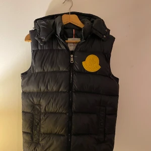 Svart dunväst från Moncler - Säljer en snygg svart dunväst från Moncler med avtagbar luva och en stor gul logga på bröstet. Den är stilren och passar bra över en hoodie eller tröja. Kom DM vid eventuella frågor eller fler bilder!