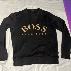 Svart tröja från Hugo Boss - Snygg svart tröja från Hugo Boss med stort BOSS-tryck i guld på framsidan. Tröjan har långa ärmar. 