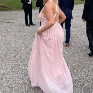Ljusrosa långklänning med pärldetaljer - Elegant ljusrosa långklänning med pärldetaljer i midjan. Klänningen har en vacker omlott-design och är axelbandslös, vilket ger en stilren look. Perfekt för ballen! Klänningen har fått lite bruns utan sol vid armhålan (med det går bort) och längst ner vid klänningen som ochså går bort. Stoleken är S men jag har M och den sitter bga