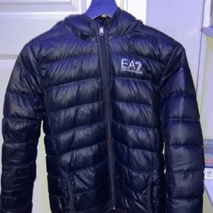 Svart pufferjacka från Emporio Armani - Säljer en svart pufferjacka från Emporio Armani EA7. Jackan har en dragkedja framtill och är quiltad för extra värme. Perfekt för kyligare dagar med sin stilrena design och bekväma passform.