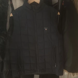 Svart quiltad väst från Gant - Snygg svart quiltad väst från Gant med dragkedja och två fickor framtill. Perfekt för lager-på-lager under kyligare dagar. Klassisk design med Gant-logga på bröstet.