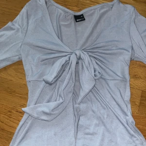 Ljusblå/grå topp från Gina Tricot - Säljer denna ljusblå/gråa slutsålda topp från Gina Tricot i storlek XS. Toppen har långa ärmar och en knytning framtill som ger en snygg detalj. Perfekt för en stilren look. Toppen är aldrig använd och det är därför jag säljer den, dock är lappen bortklippt 