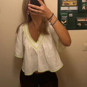 Vit blus med gröna detaljer - Säljer denna bohemiska blus med gröna broderade detaljer runt v-ringningen och ärmsluten. Blusen har en lös passform och korta ärmar, perfekt för varma dagar. Den är både stilren och bekväm. 