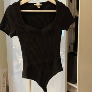 Svart ribbad bodysuit från H&M - Säljer en stilren svart ribbad bodysuit från H&M. Den har en hjärtformad urringning och korta ärmar, perfekt för en chic look. Passar bra till både jeans och kjol. Ett måste i garderoben!