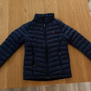 Mörkblå pufferjacka från Polo Ralph Lauren - Säljer en snygg mörkblå pufferjacka från Polo Ralph Lauren. Jackan har en dragkedja framtill och två praktiska fickor med dragkedjor. Den är dekorerad med det ikoniska röda Polo-logotypen på bröstet. Perfekt för kyligare dagar! Jackan är i storlek 8 i barn så den borde passa en person som är runt 150 - 155 cm lång!