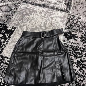 Svart skinnkjol från Pull&Bear - Snygg svart skinnkjol från Pull&Bear i storlek XS. Kjolen har en stilren design med ett bälte i midjan och en slits framtill. Perfekt för att ge en edgy touch till din outfit.