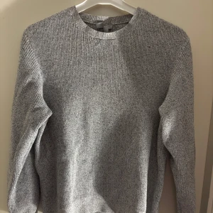 Grå stickad tröja från COS - Säljer en stilren grå stickad tröja från COS. Perfekt för kyligare dagar med sin mjuka och bekväma passform. Tröjan har långa ärmar och en klassisk rund halsringning.