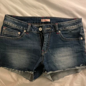 Blå jeansshorts med slitna detaljer - Snygga blå jeansshorts från Noisy May med slitna kanter och klassisk femficksdesign. Perfekta för en avslappnad stil med knapp och dragkedja framtill. Passar utmärkt till sommarens alla äventyr!