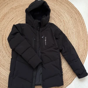 Svart dunjacka från Sail Racing - Snygg svart dunjacka från Sail Racing med dragkedja och huva. Jackan har långa ärmar och flera praktiska fickor med dragkedjor. Perfekt för kyliga dagar. Storlek 160 funkar som xs eller s