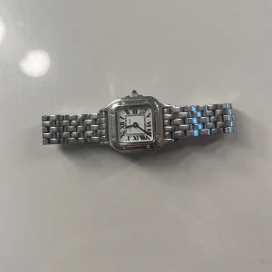 Silverfärgad klocka med länkarmband - Elegant klocka i silver med länkarmband och vit urtavla. Klockan har romerska siffror och blå visare, vilket ger en klassisk look. Perfekt för den som gillar stilrena accessoarer.