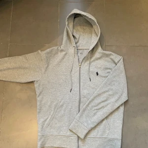 Grå hoodie från Polo Ralph Lauren - Säljer en stilren grå hoodie från Polo Ralph Lauren med dragkedja. Tröjan har inga tecken på användning! Mitt pris: 649 kr men kan diskuteras. Skriv vid behov av fler bilder eller vid minsta fundering! Nypris:1499 kr