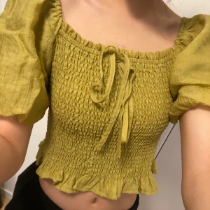 Olivgrön smockad topp - Säljer en olivgrön smockad topp med puffiga korta ärmar och volangdetaljer. Toppen har en söt knytning framtill och är croppad för en trendig look. Perfekt för varmare dagar! hör av er för frågor💞💞