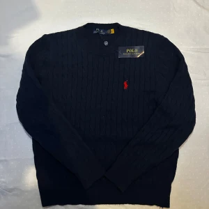 Mörkblå stickad tröja från Ralph Lauren - Säljer en stilren mörkblå kabelstickad tröja från Ralph Lauren med det ikoniska röda logotypbroderiet på bröstet. Tröjan har långa ärmar och en klassisk rund halsringning. Perfekt för en elegant och avslappnad look.