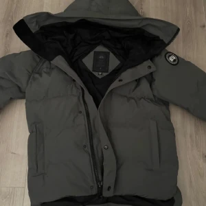Grå dunjacka från Canada Goose - Säljer en grå dunjacka från Canada Goose med huva och dragkedja. Jackan har en klassisk design med en logotyp på ärmen. Perfekt för kalla dagar.