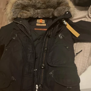 Para jumper svart parkajacka  - Säljer en svart parkajacka från Parajumpers med pälsfodrad huva och dragkedja. Jackan har flera fickor och en gul detalj vid halsen. Perfekt för kalla dagar.