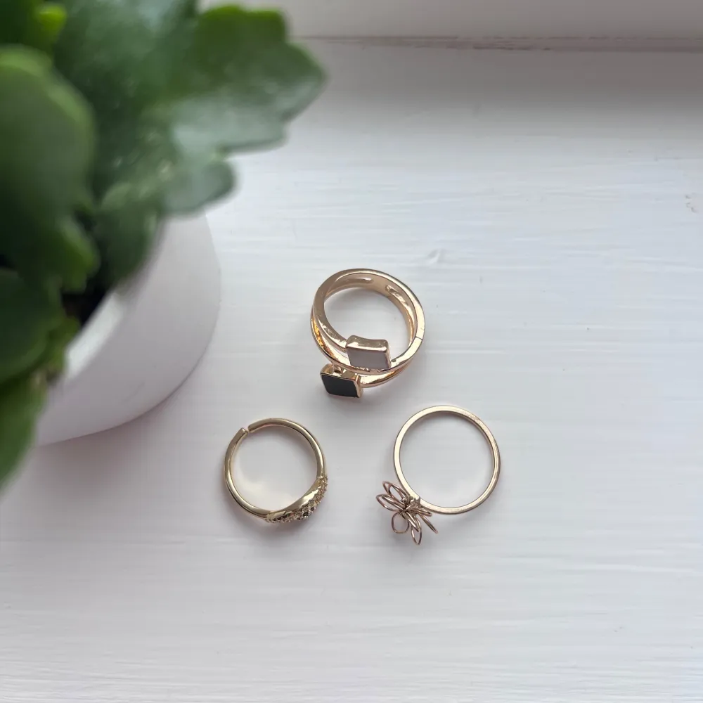 Snyggt set med tre guldringar. En ring har en svart och vit fyrkantig detalj, en annan har en blomformad design, och den tredje har en ögonformad detalj med små stenar. Perfekt för att mixa och matcha! Jag lägger ringarna i plastfickan som finns på sista bilden, så de inte utsätts för skada under leverans.. Asusteet.