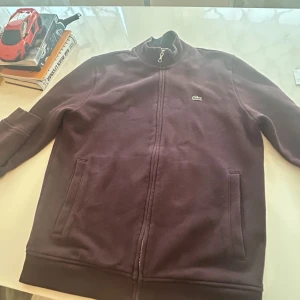 Zip tröja från Lacoste - Säljer en snygg mörklila tröja från Lacoste med dragkedja och broderad logga på bröstet. Tröjan har långa ärmar och en hög krage. Perfekt för en stilren look!