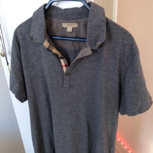 Grå pikétröja från Burberry - Säljer en stilren grå pikétröja från Burberry Brit i 100% bomull. Tröjan har korta ärmar och en klassisk krage med knappar. Perfekt för en avslappnad men ändå elegant look.
