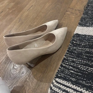 Beige pumps från Tamaris - Snygga beige pumps från Tamaris med spetsig tå och klack. Perfekta för en elegant look. Tillverkade i ett stilrent material som ger en sofistikerad touch.