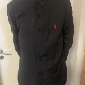 Svart skjorta från Ralph Lauren - Snygg svart skjorta från Ralph Lauren med röd broderad logga på bröstet. Skjortan har långa ärmar och knappar framtill. Perfekt för en stilren look.