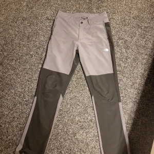 Grå och svarta byxor från The North Face - Snygga och funktionella byxor från The North Face i grått och svart. Byxorna har en normal passform med dragkedja och knapp framtill. Perfekta för utomhusaktiviteter med slitstarkt material och logotypdetalj på benet.