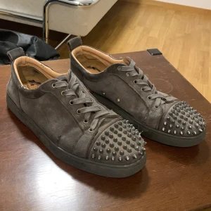 Gråa Christian Louboutin skor - Snygga gråa Christian louboutins i storlek 44. Skorna är ganska så slitna då sulan har börjat slipas bort, 2 nitar har ramlat av och de är ett litet hål på sidan av höger sko som man ser på första bilden. Har box, dustbag, extra snören och bild på kvittot.! Skriv vid frågor och kom gärna med prisförslag!